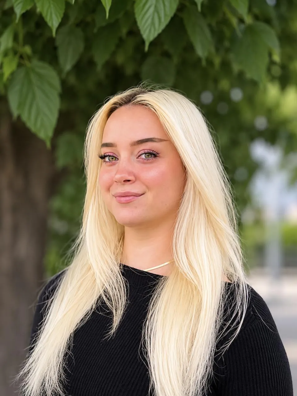 A woman with long blonde hair wearing a black top stands outdoors in front of a tree with green leaves.