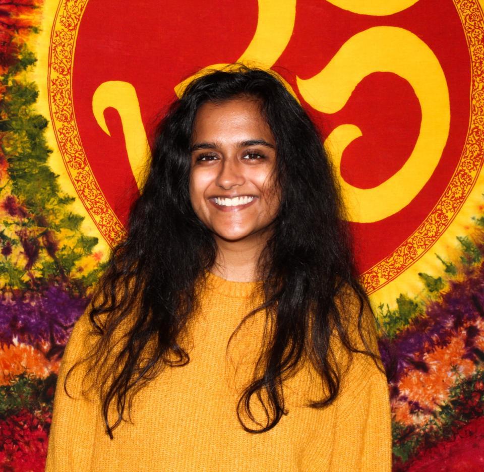 A person with long dark hair and a yellow sweater smiles in front of a vibrant tapestry with red and yellow patterns.