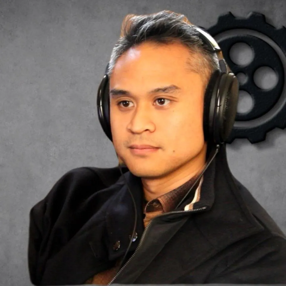A person with short, dark hair wearing large headphones and a dark jacket sits in front of a gray background with a gear symbol.