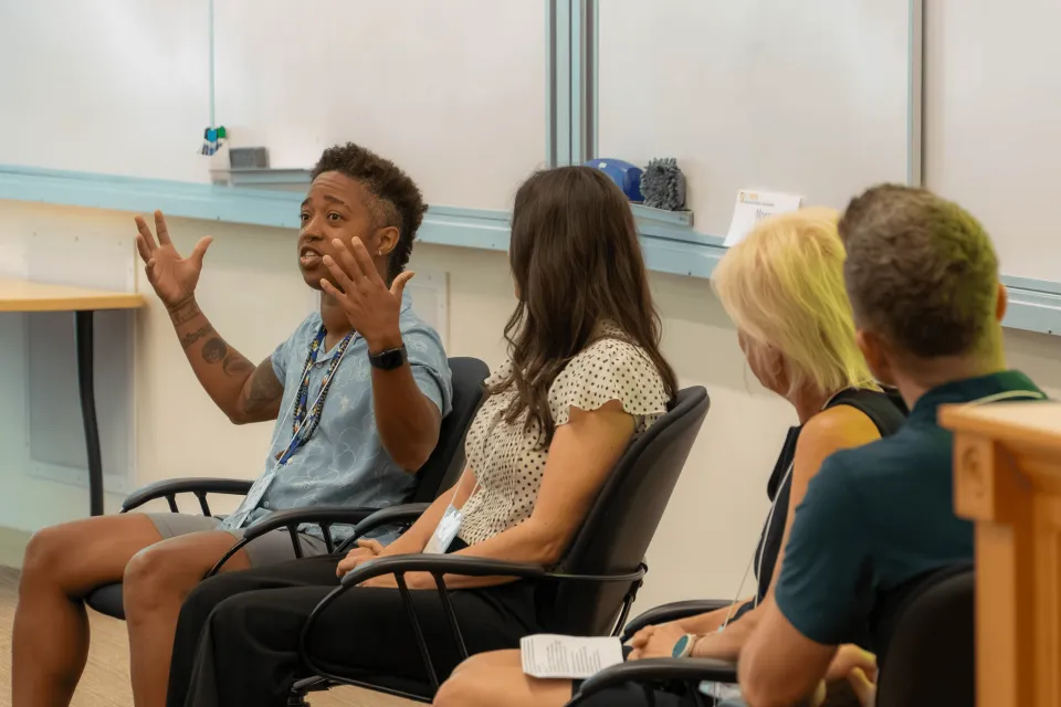 Four people sit in chairs in a classroom setting; one person gestures with both hands while speaking, and the others listen attentively.