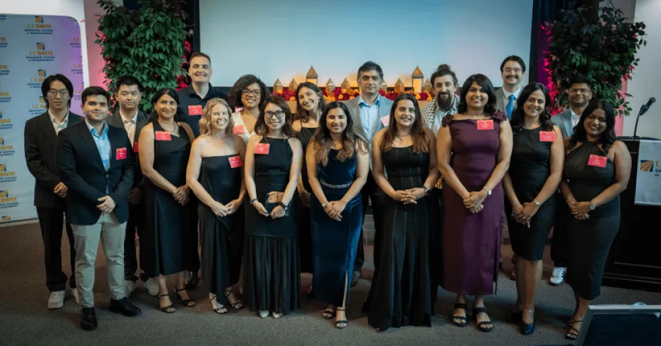 A group of nineteen people, dressed in semi-formal attire, stand in two rows posing for a photo indoors at an event.