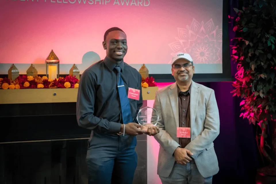 Two men stand indoors smiling at the camera; one holds a clear award plaque. Both wear name tags and are positioned in front of a decorated stage display.