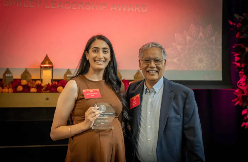 Two people stand together indoors. The woman holds an award and smiles at the camera. A sign behind them reads "Leadership Award." The setting features decorative lights and floral elements.