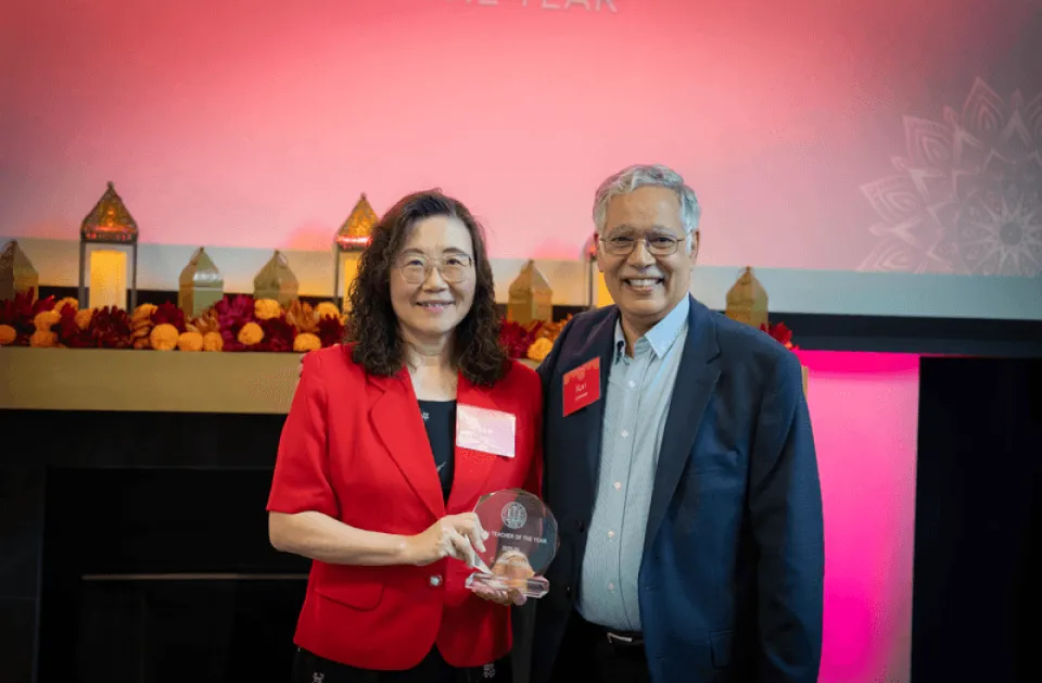 Two people stand smiling in front of a screen displaying "C. Janie Chang Teacher of the Year." The woman holds an award and both are dressed in business attire.