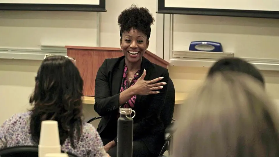 A woman in business attire smiles and gestures while speaking to a group in a classroom or conference setting.