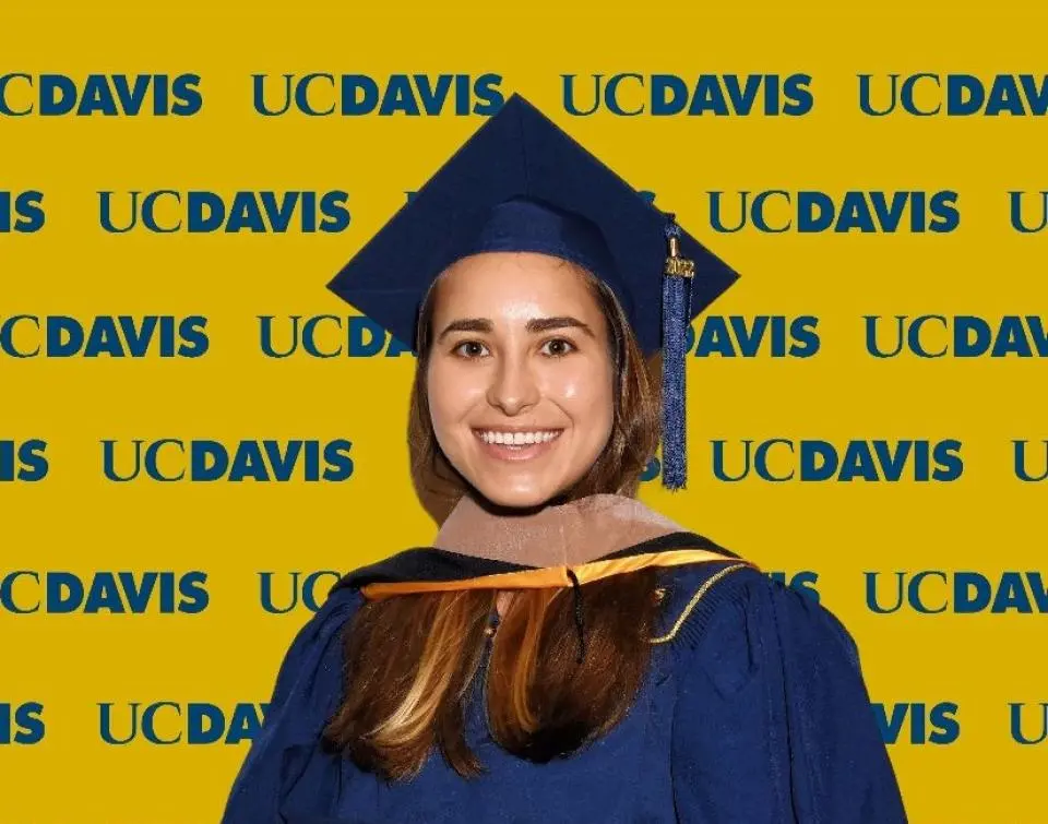 Maria Ahumada wearing a blue graduation cap and gown stands in front of a yellow UC Davis backdrop with repeated logos.