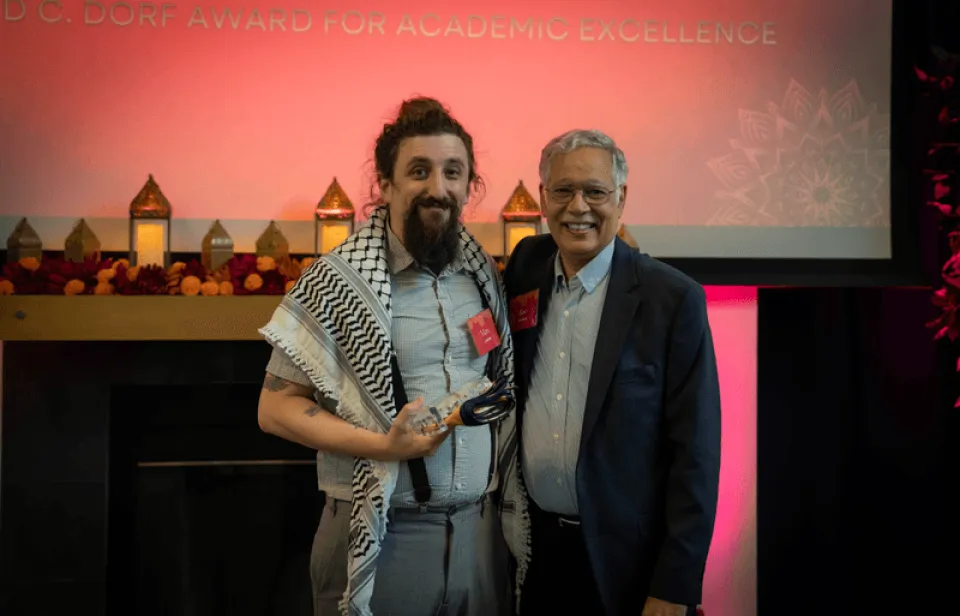 Two men stand together smiling; one holds a glass award. A screen behind them displays "D.C. Dorf Award for Academic Excellence." Decorative lanterns are arranged on a mantel.
