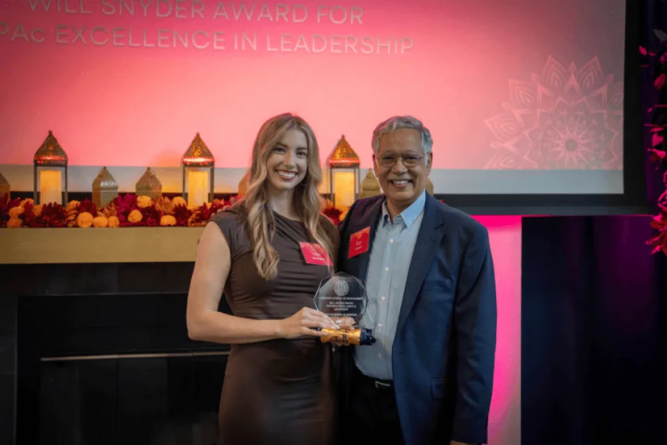 A woman and a man stand together indoors, smiling. The woman holds an award. A screen behind them reads "EXCELLENCE IN LEADERSHIP." Decorative lanterns are on a mantel.