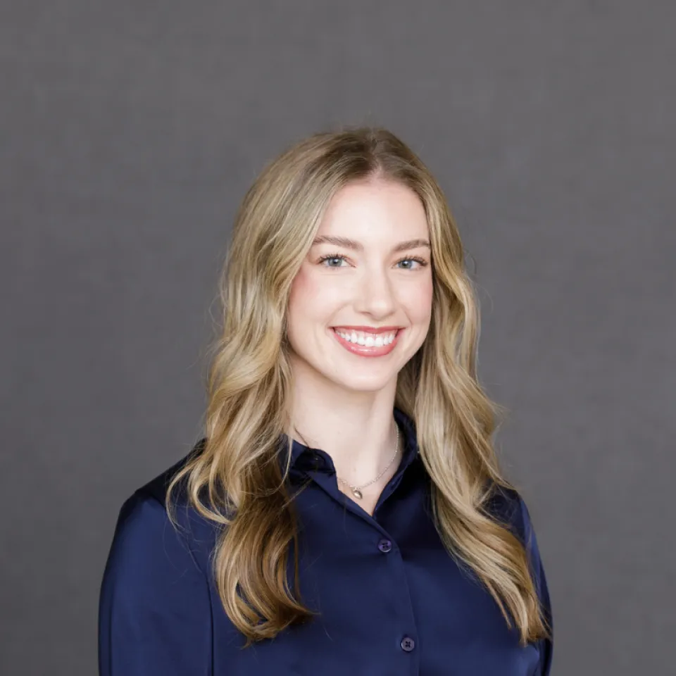 A young woman with long, wavy blonde hair wearing a dark blue shirt, smiling in front of a plain gray background.