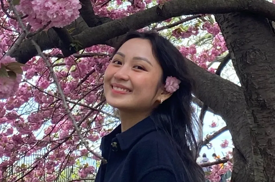 A woman with long dark hair and a flower behind her ear smiles while standing under a tree with pink blossoms.