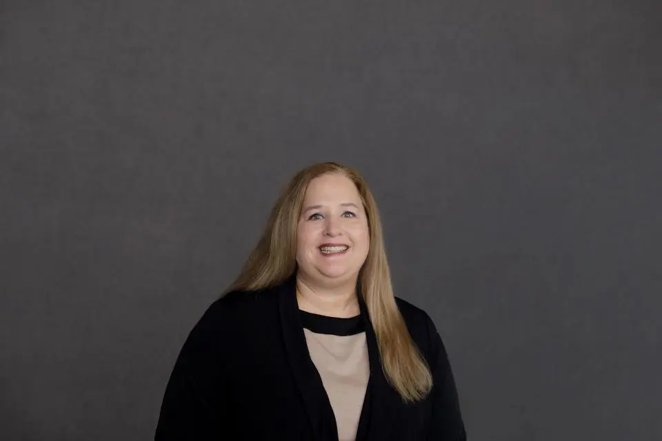 A woman with long blonde hair, wearing a beige top and black cardigan, stands smiling in front of a plain gray background.