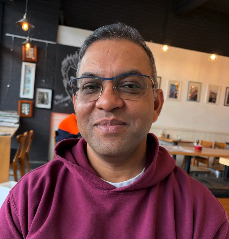 A man wearing glasses and a maroon hoodie sits in a cafe, smiling at the camera. Framed pictures and wood furniture are visible in the background.