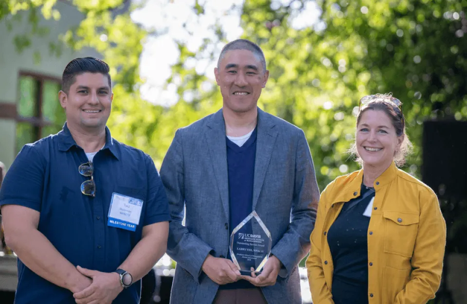 Three people stand outdoors smiling; the person in the center holds a clear award plaque. Trees and sunlight are visible in the background.