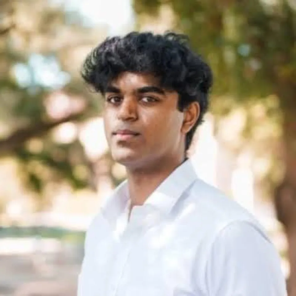 Anirudh Murugesan wearing a white shirt stands outdoors, with trees and sunlight blurred in the background.
