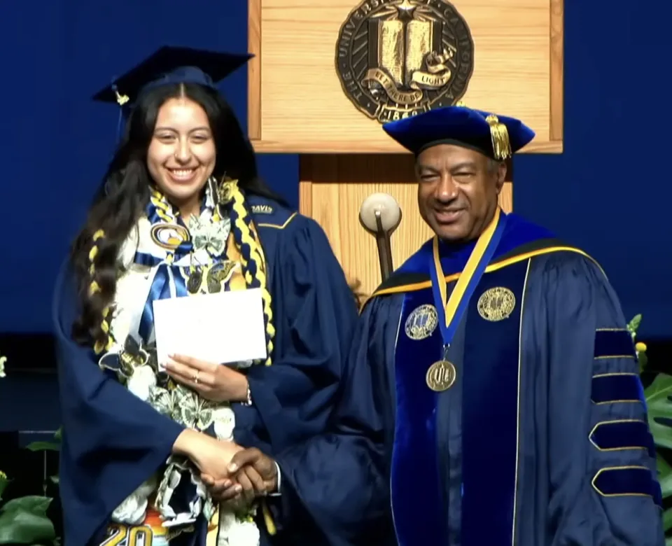 A graduate in a cap and gown shakes hands with a university official in academic regalia on stage at a commencement ceremony.
