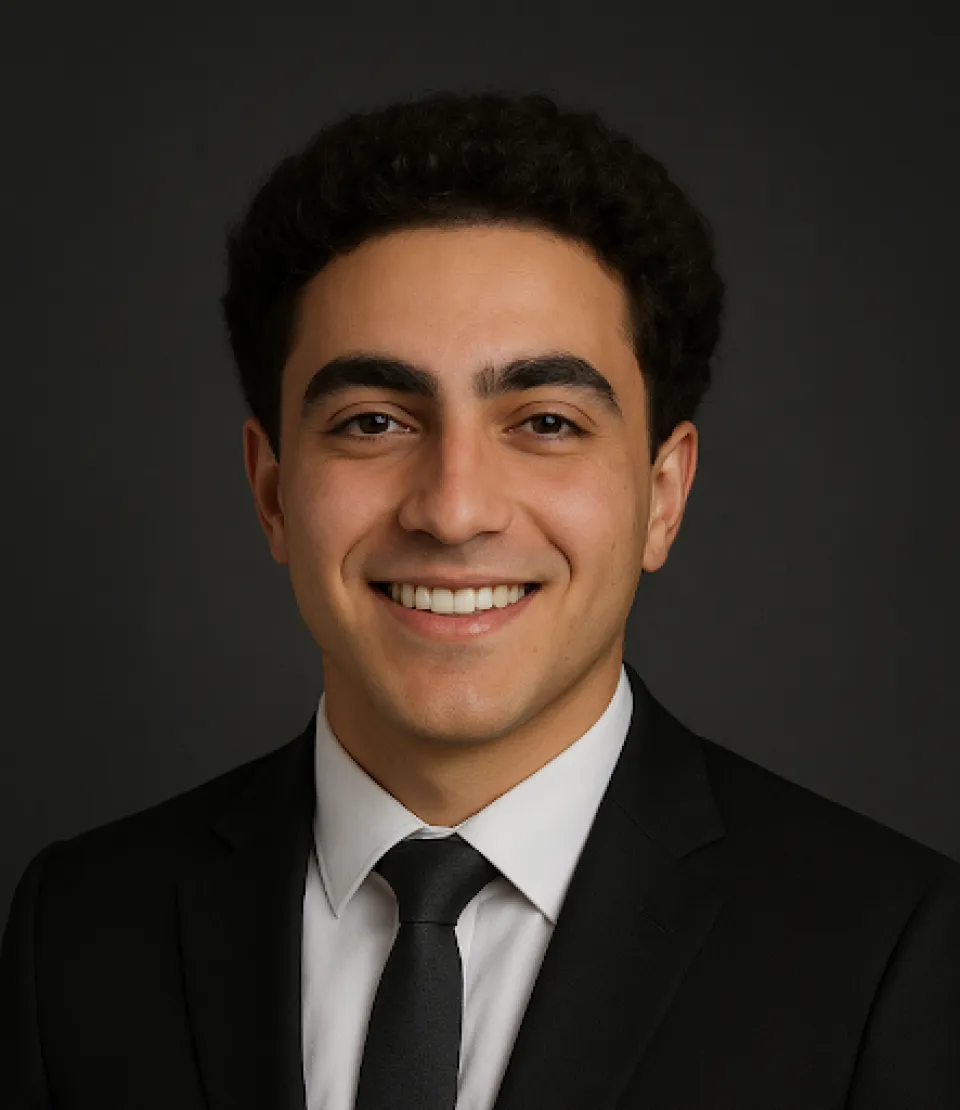 Mark Soliman, wearing a black suit, white shirt, and black tie, smiling in front of a plain dark background.