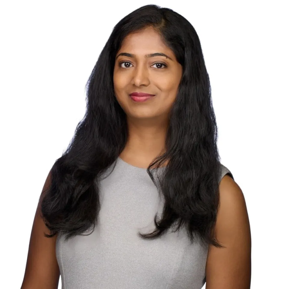 Smita Madam wearing a sleeveless light gray top stands in front of a plain white background, looking at the camera and smiling slightly.