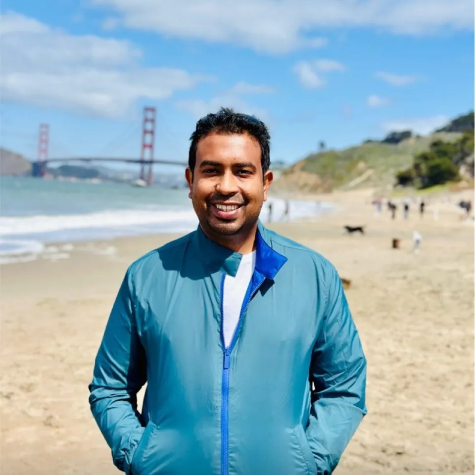 Man in a blue jacket stands on a sandy beach with the Golden Gate Bridge and blue sky in the background.