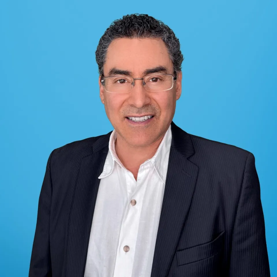Benigno Salazar wearing a dark suit jacket over a white shirt stands smiling in front of a solid blue background.