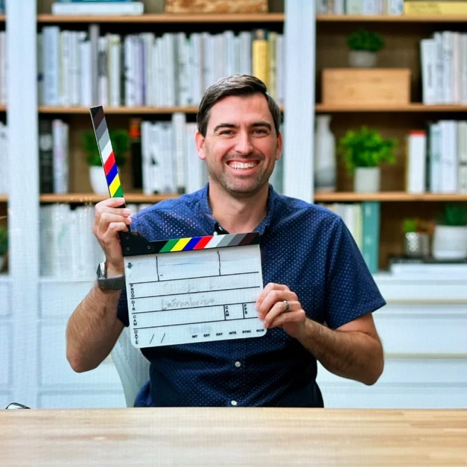Man sitting at a desk, smiling and holding a film clapperboard in front of a bookshelf with books and plants.