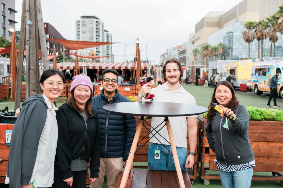 Five people stand smiling around a high outdoor table at a market event, with food trucks, palm trees, and buildings in the background.