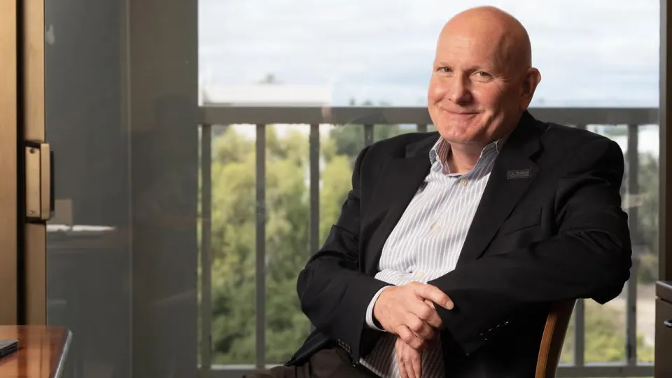 A bald man in a suit jacket sits at a desk, smiling, with trees and a cloudy sky visible through a window behind him.