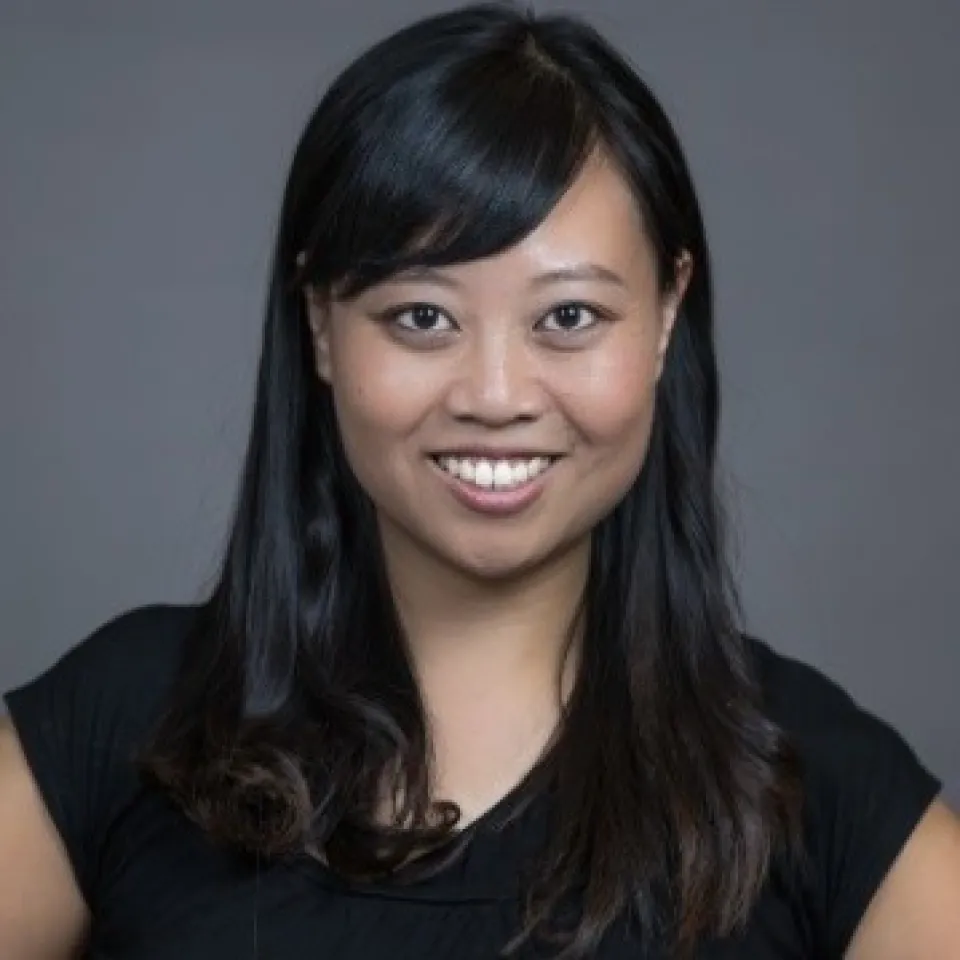 Woman with long black hair smiling, wearing a black top, posing in front of a plain gray background.