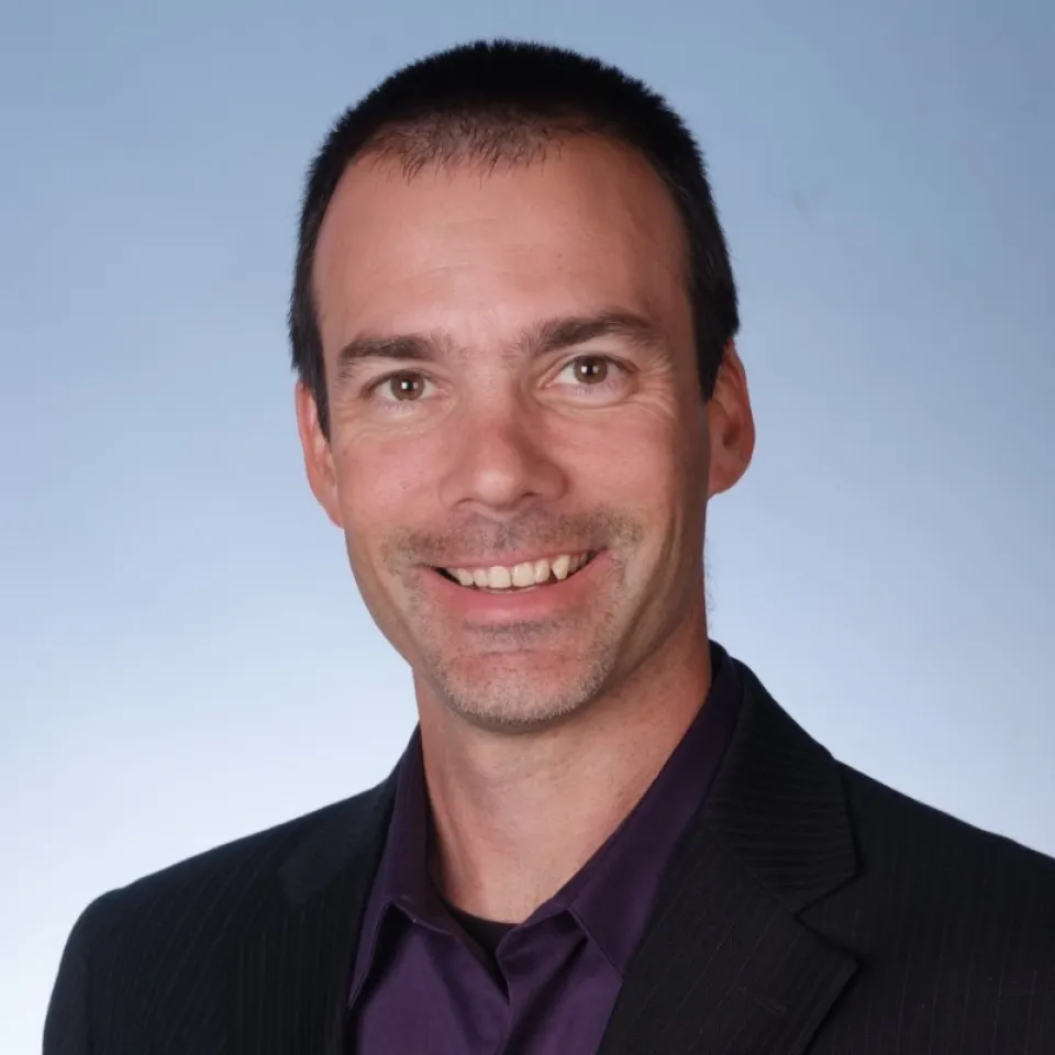 Nate Whaley wearing a black suit jacket and dark purple shirt, smiling in front of a plain light blue background.