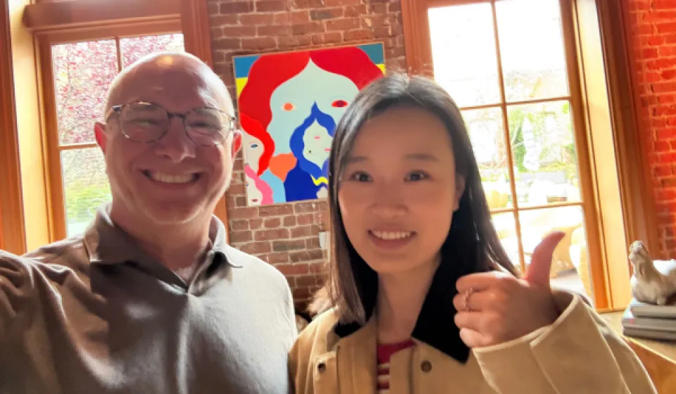Two people smile at the camera indoors; one gives a thumbs up. A colorful abstract painting and large windows with a garden view are in the background.