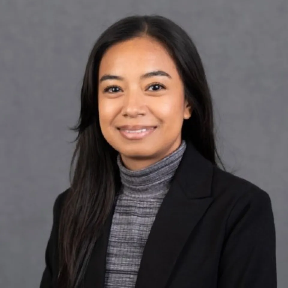 Savanna De La Paz wearing a black blazer and gray turtleneck, smiling in front of a plain gray background.