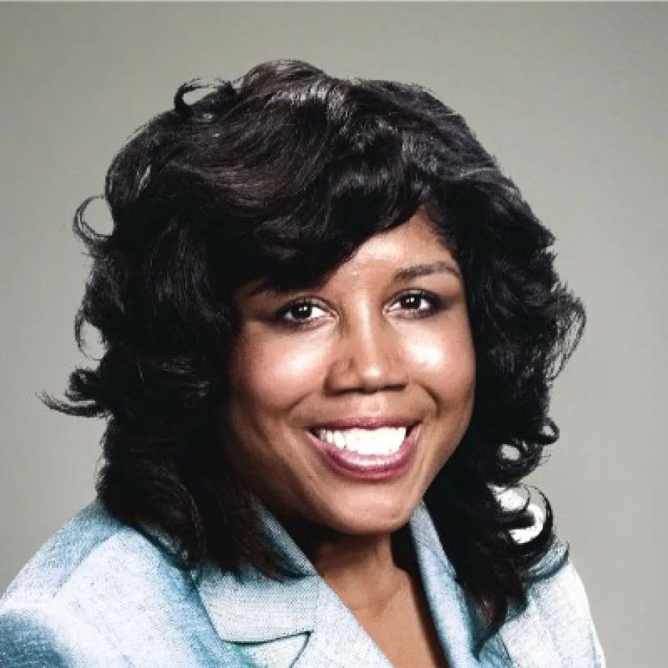 Smiling woman with dark curly hair wearing a light-colored blazer poses in front of a plain, neutral background.