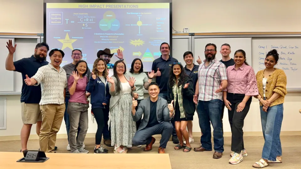 A large group of people posing in a classroom in front of a presentation screen and a whiteboard with handwritten notes.