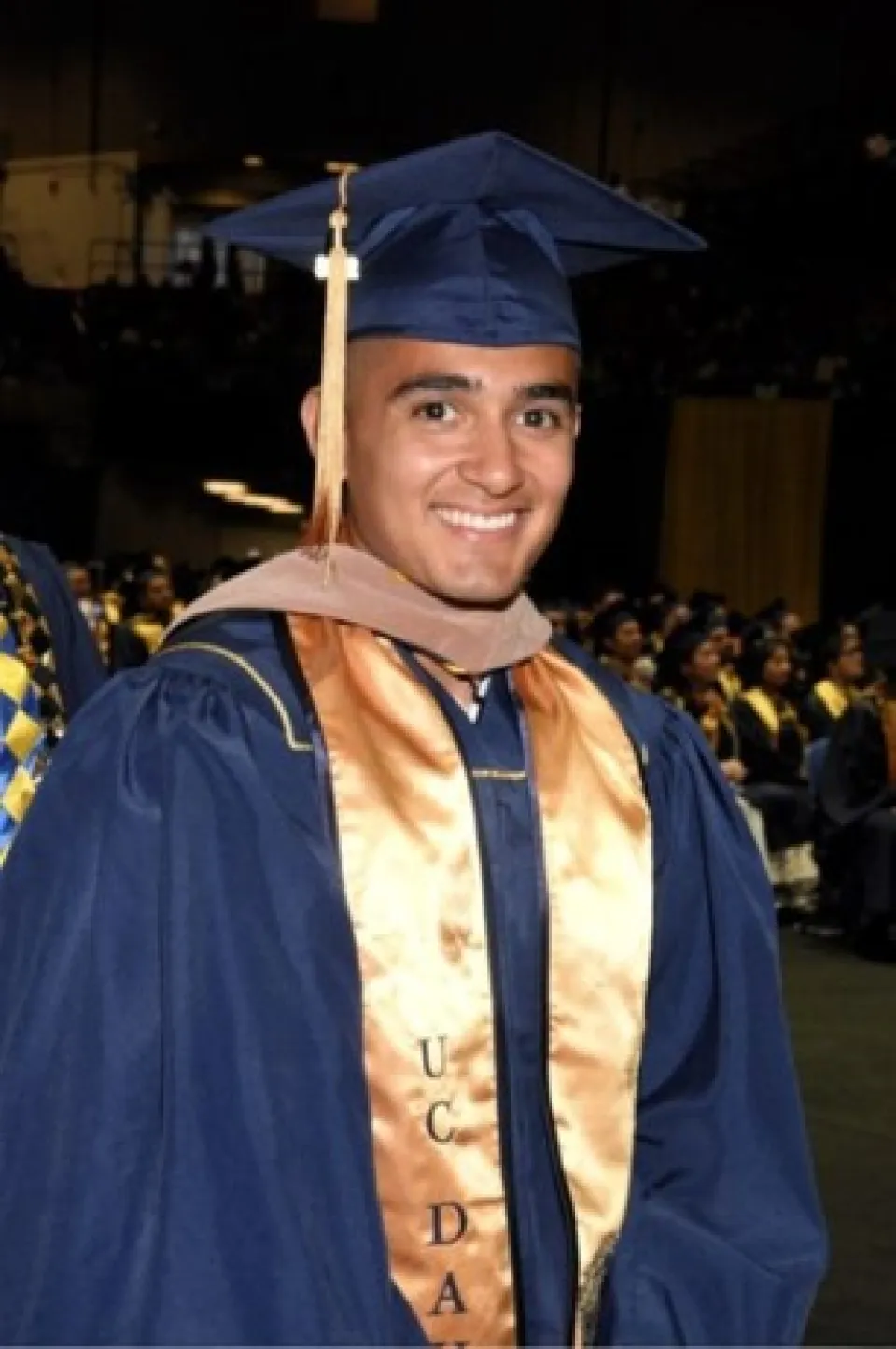 A smiling graduate in a blue cap and gown with a gold stole at a commencement ceremony.