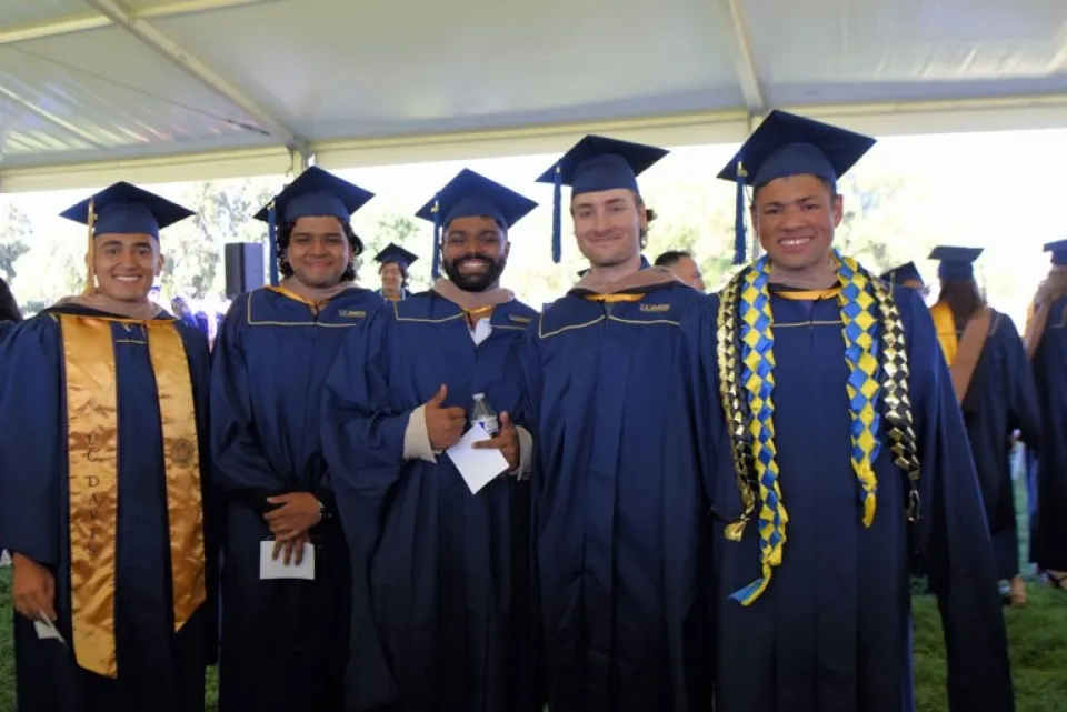 Five students in UC Davis graduation caps and gowns pose together outdoors.