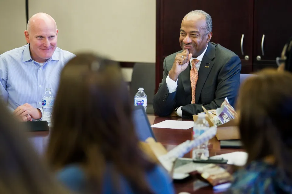 A man in a suit and a man in a button-down shirt smile at each other across a conference table during a meeting.