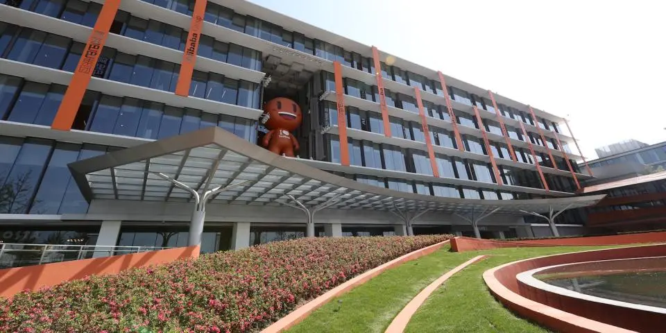 Modern office building with large glass windows and orange accents, featuring a smiling cartoon figure on an upper floor and landscaped greenery in the foreground.