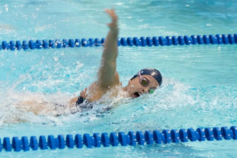Swimmer wearing goggles and a swim cap performs freestyle stroke in a lane of a swimming pool, with blue lane dividers visible.