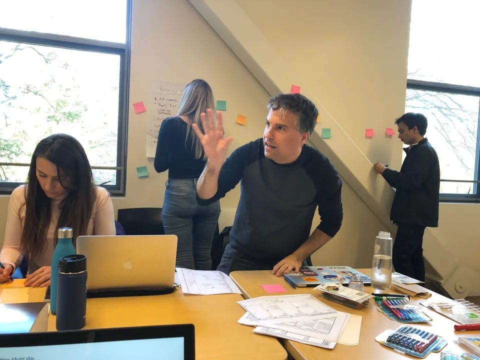 Four people work in a bright room; one gestures while speaking, two write on paper, and one writes on sticky notes on the wall. Desks hold notebooks, markers, and water bottles.