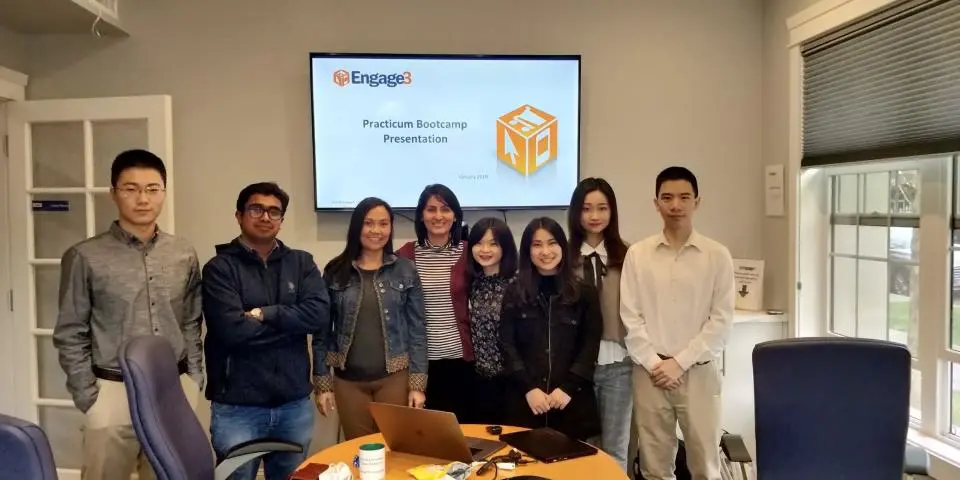 A group of eight people stands in a conference room in front of a screen displaying "Engage3 Practicum Bootcamp Presentation.