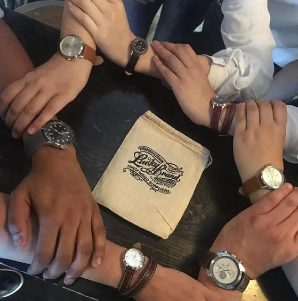 Six hands wearing different wristwatches form a circle around a Lucky Brand fabric pouch on a dark table.