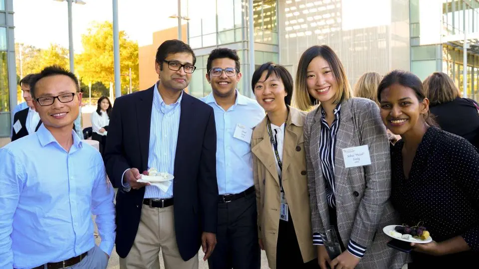 A group of six people, dressed in business attire, pose and smile at an outdoor event with a modern glass building in the background.