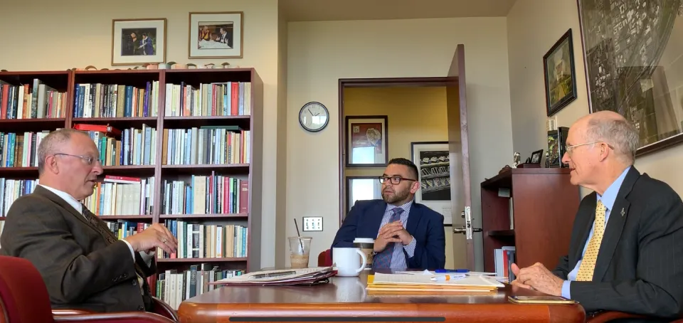 Three men in suits have a discussion in an office lined with bookshelves; one sits across from the other two at a desk with papers, a mug, and a coffee cup.
