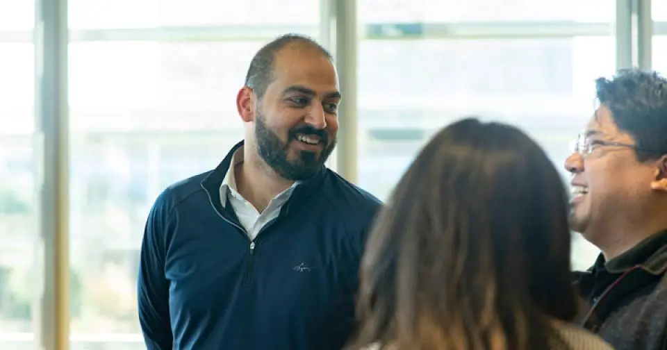 Three people stand indoors near large windows, engaged in conversation. One man in a dark pullover is smiling while talking to two others.