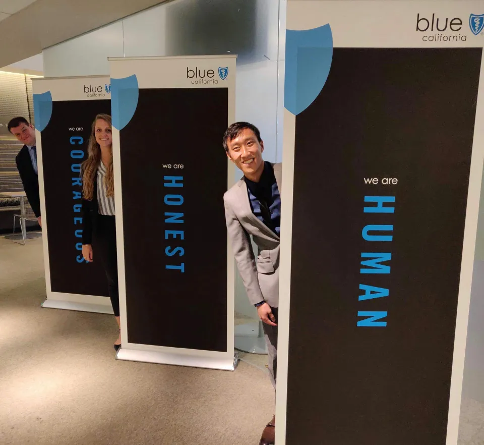 Four people in business attire stand behind three banners labeled "Courageous," "Honest," and "Human" at a Blue California office.