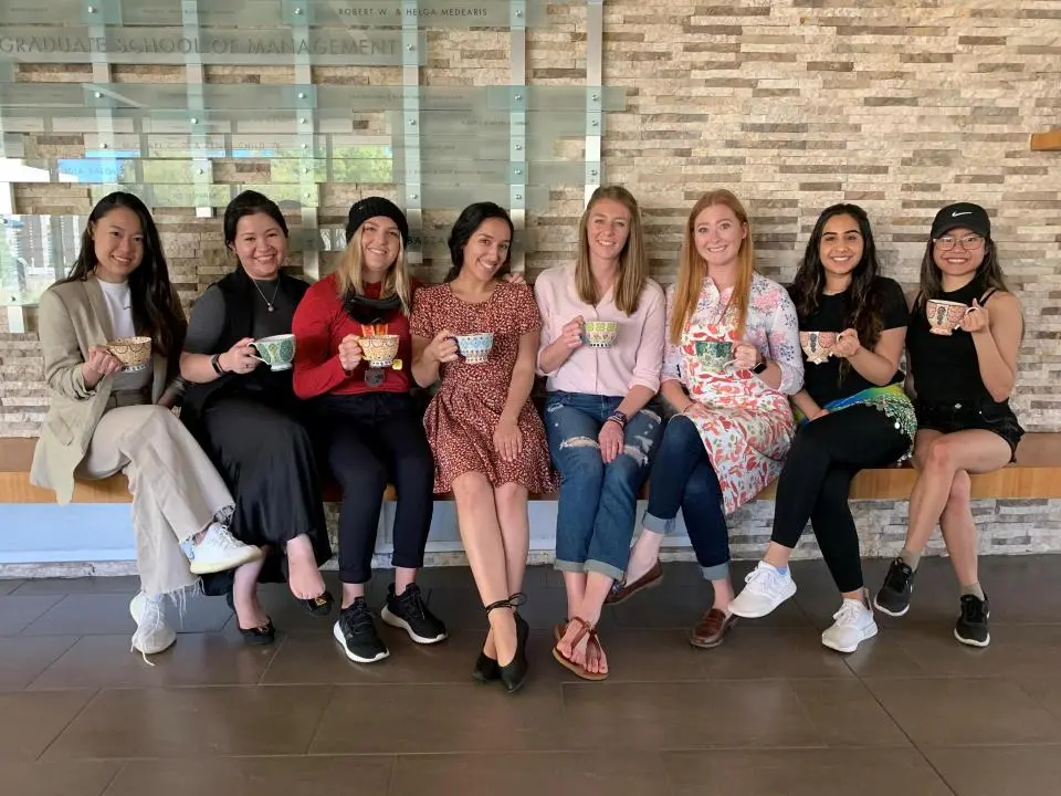 Seven women sit on a bench indoors, smiling and holding colorful mugs, with a stone wall and glass panels in the background.