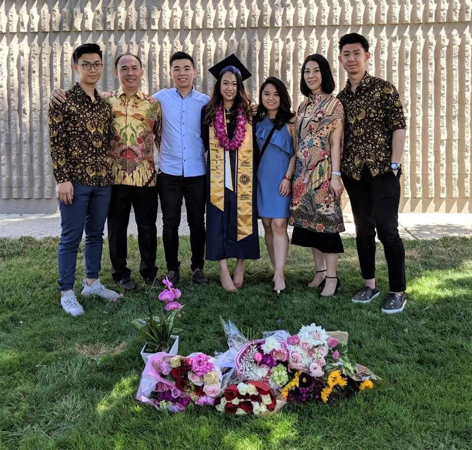 Six people stand together outdoors, with one woman in graduation attire and a lei. Flower bouquets are arranged on the grass in front of them. They pose in front of a textured wall.