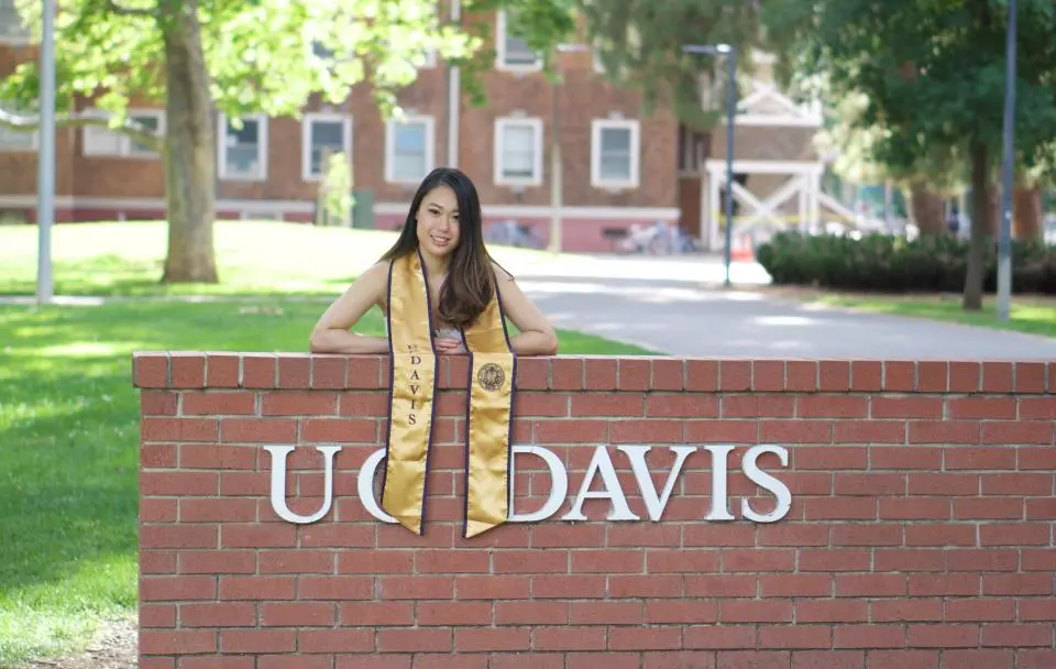 A woman in a graduation stole stands behind a red brick wall with "UC DAVIS" in white letters, with campus buildings and trees in the background.