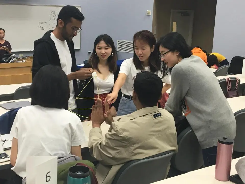 Five students work together at a classroom desk to build a tower using spaghetti sticks and marshmallows during a group activity.