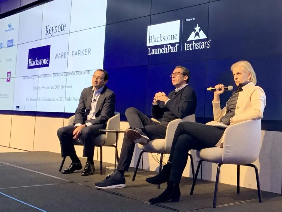 Three speakers sit on stage in front of a large screen displaying business logos and the words "Keynote Blackstone LaunchPad powered by techstars." One person speaks into a microphone.