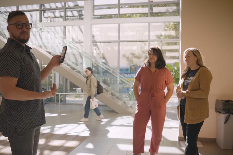 Tim McConville holds up a phone to take a photo of Espy Foft and Amy Davis standing and smiling in a sunlit indoor space with large windows and a staircase.